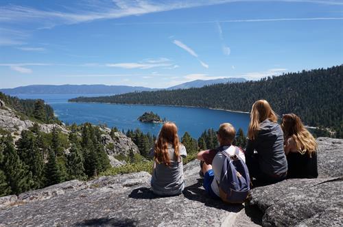 5 Young peopla are sitting on a huge rock and are watching a big lake lying below them.